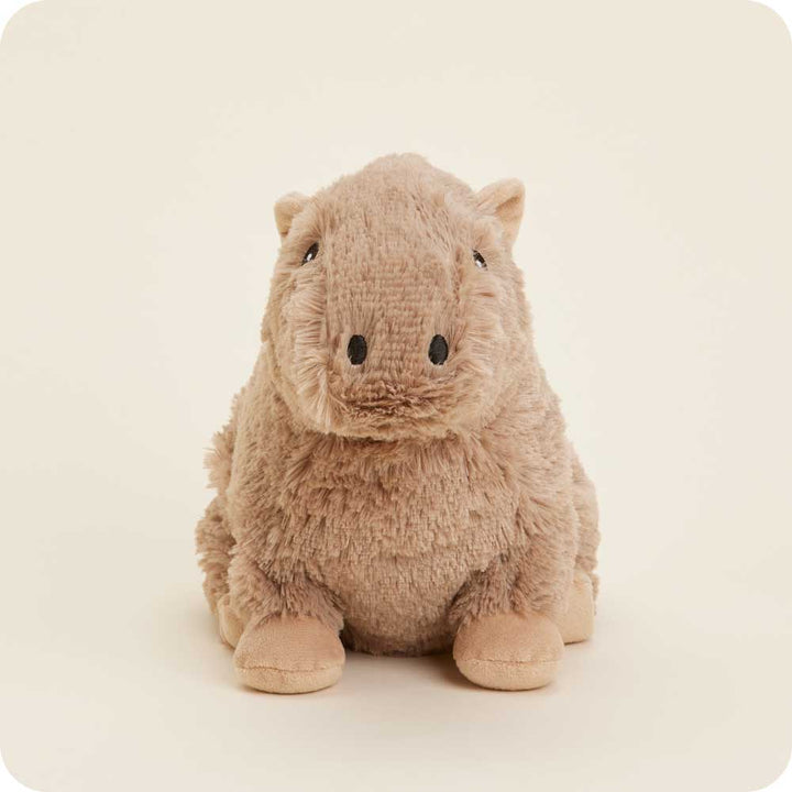 A Capybara Warmie by Warmies, with its light brown color, small black eyes, and slightly smiling expression, rests against a pale background, providing a cute and cuddly look.