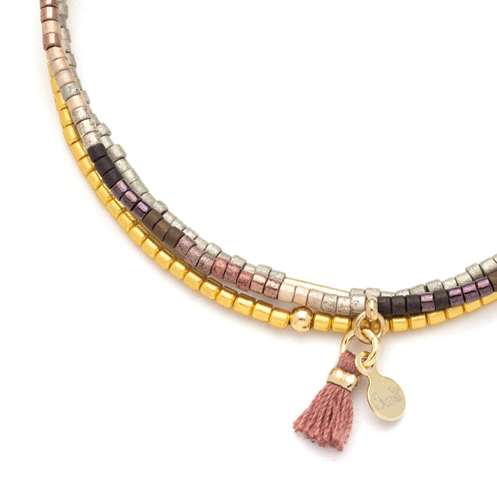 A close-up of the Bronze Multi Gold Miyuki Bracelet Trio by Scout Jewelry, featuring rows of small, square Miyuki Delica beads in metallic shades of gold, silver, bronze, and rose. The bracelet trio includes a pink tassel charm and a small gold tag hanging from it. The intricate design complements beautifully with stacking bracelets for an elegant look.