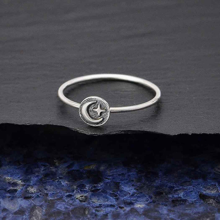 A Nina Designs s9 Raised Moon and Star Stacking ring in sterling silver, featuring a circular charm with a moon and star design, rests on a dark textured surface highlighted by a vibrant blue section underneath.