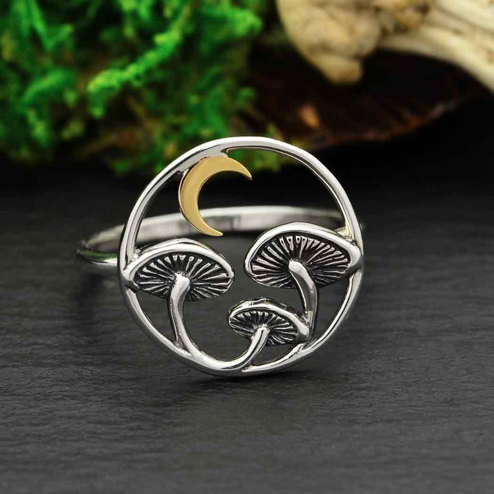 A circular design featuring two mushrooms and a bronze crescent moon on a textured dark surface with green foliage, this silver Sz7 Mushroom Ring from Nina Designs evokes the healing properties often associated with spirit medicine.