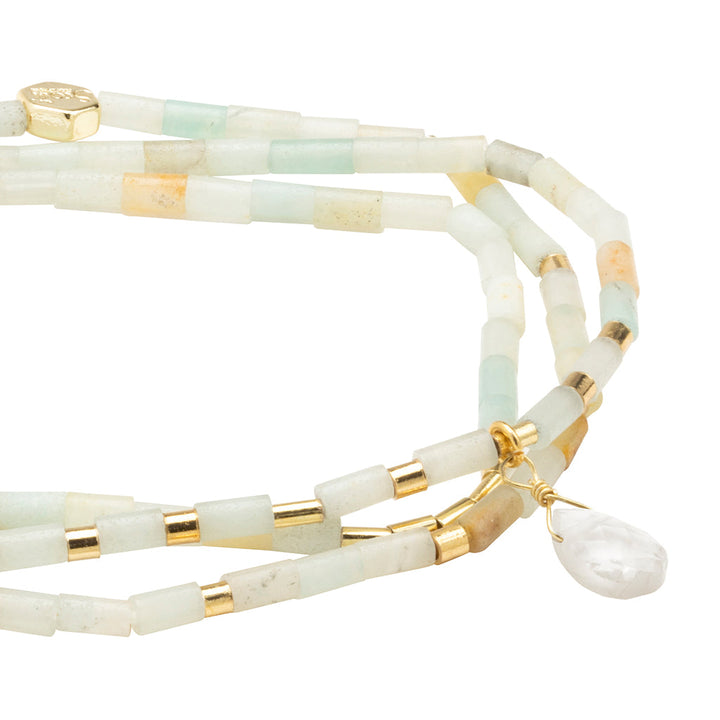 A close-up image of the Scout Jewelry Courage Amazonite Gold Teardrop Wrap, featuring a multi-strand bracelet composed of rectangular semi-precious stone beads in pastel colors, primarily light green, with gold accents. A small teardrop-shaped white pendant hangs from the bracelet.