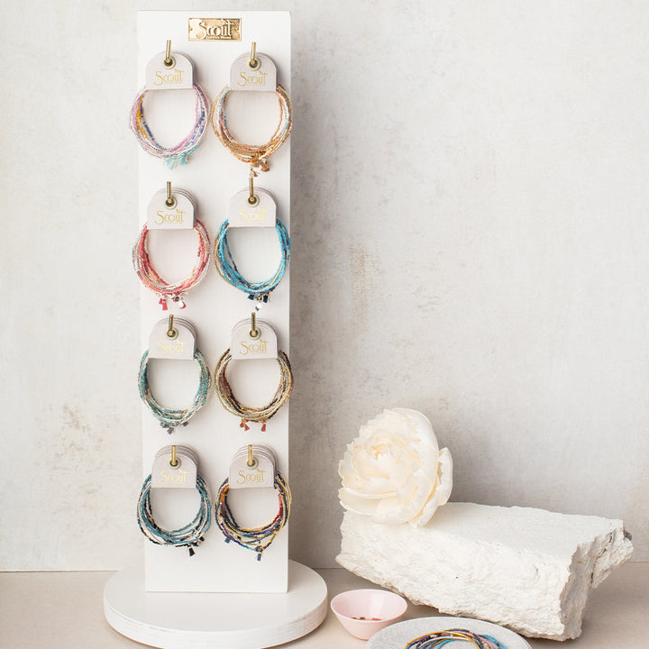 A white display stand beautifully showcases multiple sets of the Bright Multi Silver Miyuki Bracelet Trio, ideal for stacking. Each bracelet is tagged with "Scout Jewelry," and their vibrant hues are reminiscent of Miyuki Delica beads. Adding to the charm, a white flower and a light pink dish rest on a textured surface nearby.