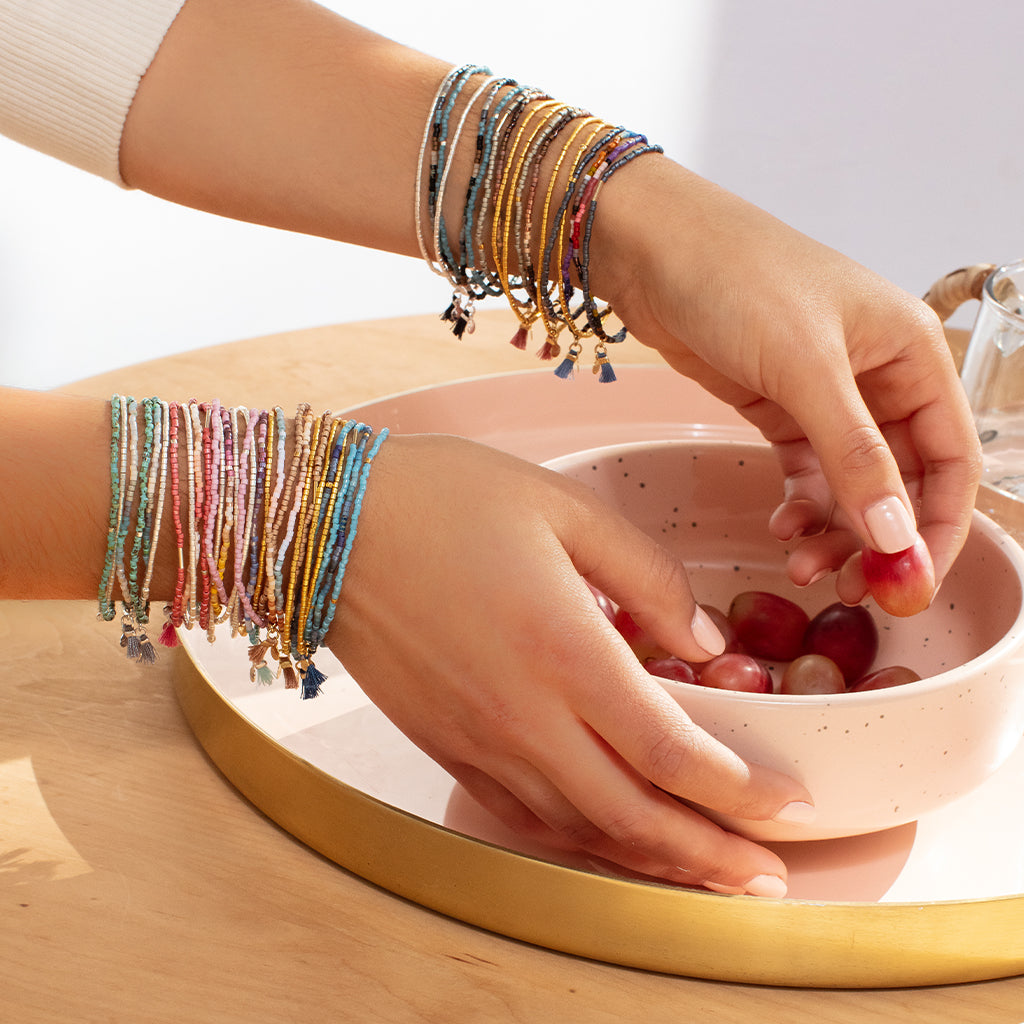 A person embellished with the Bright Multi Silver Miyuki Bracelet Trio from Scout Jewelry holds a pink bowl filled with grapes. The bowl is placed on a round, golden tray upon a wooden surface, gently lit by sunlight.