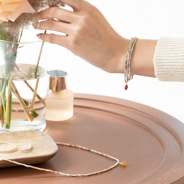 A hand adorned with a beaded bracelet featuring semi-precious stone beads reaches toward a glass vase filled with flowers on a round table. Next to the vase are a perfume bottle, a wooden tray with glasses, and the Magic Labradorite Gold Teardrop Wrap necklace by Scout Jewelry, creating a soft, elegant ambiance.