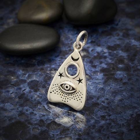 The Silver Ouija Planchette Charm with All Seeing Eye by Nina Designs is a silver pendant shaped like a planchette, echoing the mystique of an Ouija board piece. It features an eye design with a crescent moon and stars above, resting on a textured dark blue surface next to smooth black stones, evoking the enigmatic aura of seances and communicating with the deceased.