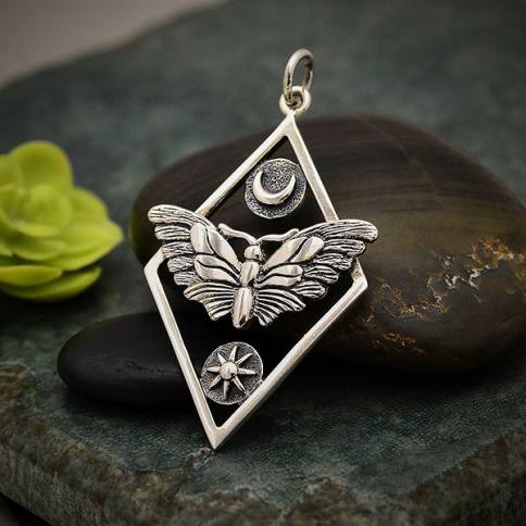 The Silver Geometric Moth Charm with Sun and Moon from Nina Designs features a detailed butterfly with outspread wings, a crescent moon above it, and a sun below it, all enclosed in a diamond-shaped frame. The charm is set against a dark, textured stone background with a small green succulent to the side, evoking psychic awareness.