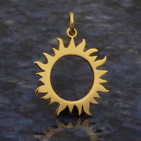 Nina Designs' Bronze Eclipse Sun Charm, featuring a gold sun shape with a hollow center and stylized pointed rays, elegantly rests on a textured dark surface, capturing the celestial mystery.