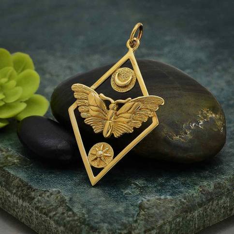 The Bronze Geometric Moth Charm with Sun and Moon from Nina Designs embodies intuition and passionate pursuits. This exquisite pendant features a moth, a sun, and a crescent moon elegantly framed within a geometric design. The elements are gracefully arranged on a stone surface, accompanied by a small green plant in the background—a delicate balance of nature and artistry.