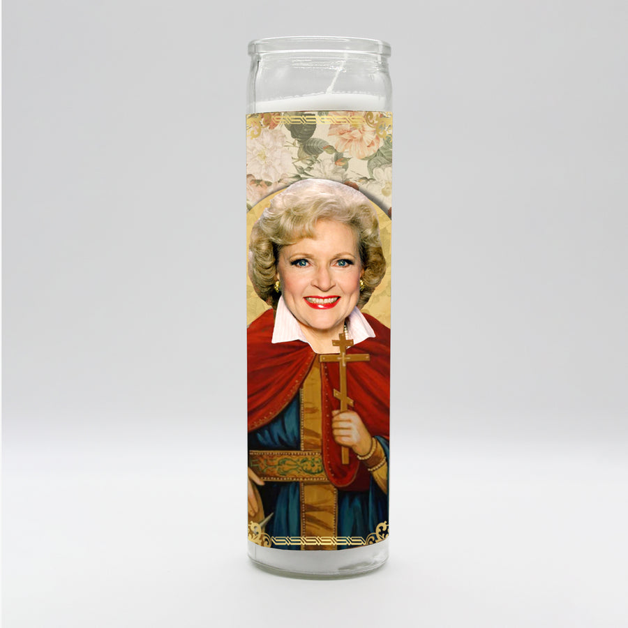 The tall, unscented "Betty White Candle" from BOBBYK BOUTIQUE features a depiction of a woman with short blonde hair, smiling widely. Dressed in a decorative robe and holding a cross, she bears a striking resemblance to Betty White. The candle's floral background enhances the saintly, religious iconography theme, making it an ideal addition to any celebrity shrine.