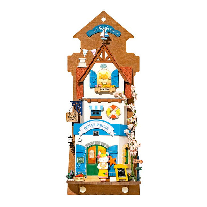 Introducing the DIY Miniature Wall Hanging Kit Island Dream Villa by Hands Craft—a charming wooden piece crafted in the shape of a vibrant house with an "Ocean House" sign. This perfect gift idea features a small lifebuoy, floral accents, and miniature sea creature figurines. Ideal for crafters and collectors, this coastal-themed masterpiece boasts stunning blue, orange, and brown hues.
