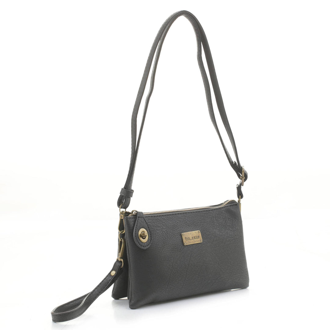 The Ella Crossbody in Black by Liz Soto Handbags is a sleek small leather bag featuring an adjustable strap, a gold zipper, and a small front pocket. It has a gold nameplate with engraved text on the front and is photographed against a plain white background.