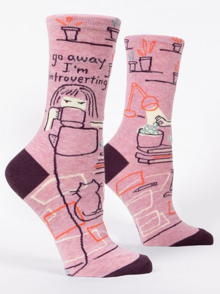 Here's a pair of "Go Away, I'm Introverting" socks by Blue Q, featuring simple illustrations of a person holding a book and drinking from a mug. The design includes shelves with plant pots and books, while the toe and heel sections are in a darker pink shade.