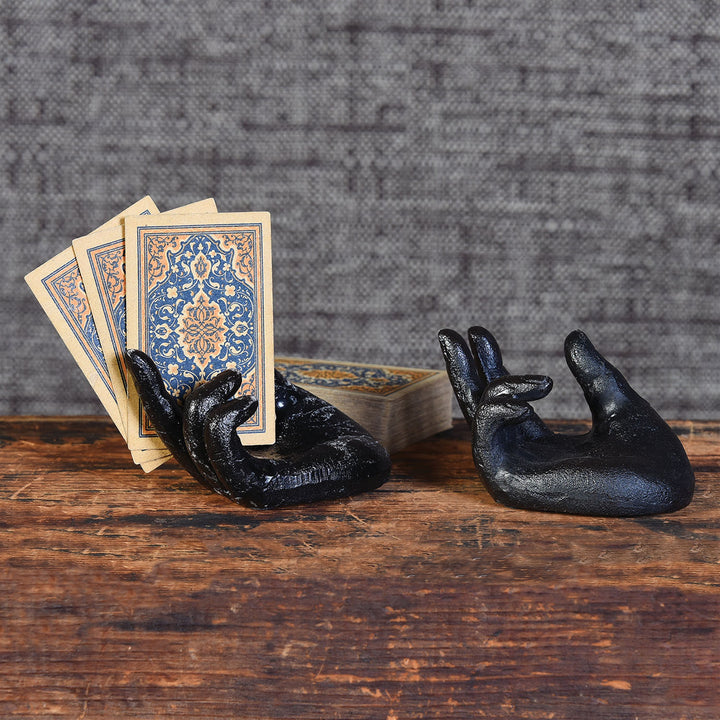 Two Single Hand Card Holders in Antique Black from HomArt rest on a wooden surface; one, resembling a zen hand, holds three ornate playing cards with intricate blue and gold designs, while the other remains empty. A stack of similar cards sits sturdily in the background against a textured gray wall.