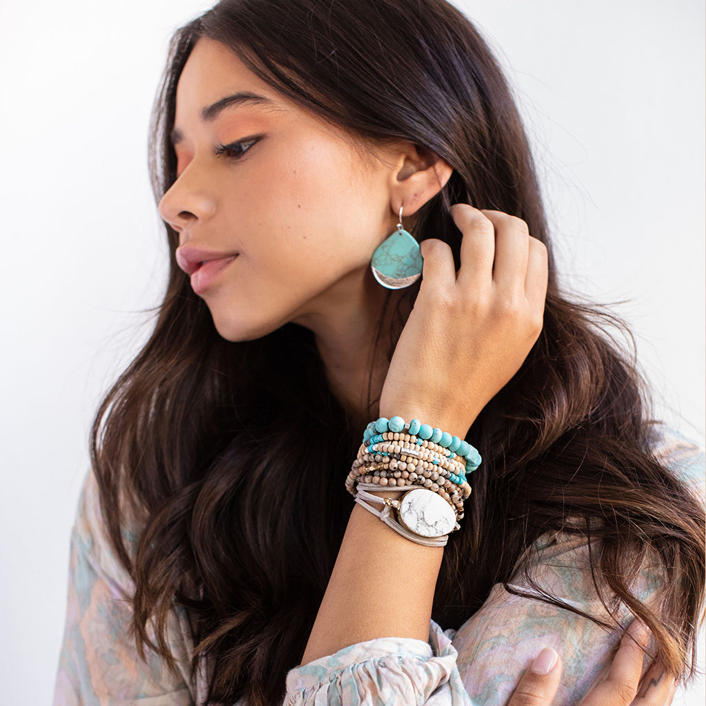 A young woman with long dark hair is wearing Scout Jewelry's Dipped Teardrop Earring in Turquoise and Silver alongside multiple beaded bracelets. She rests her hand on her neck, showcasing a white-faced watch among the bracelets, while her expression remains serene against a light background.