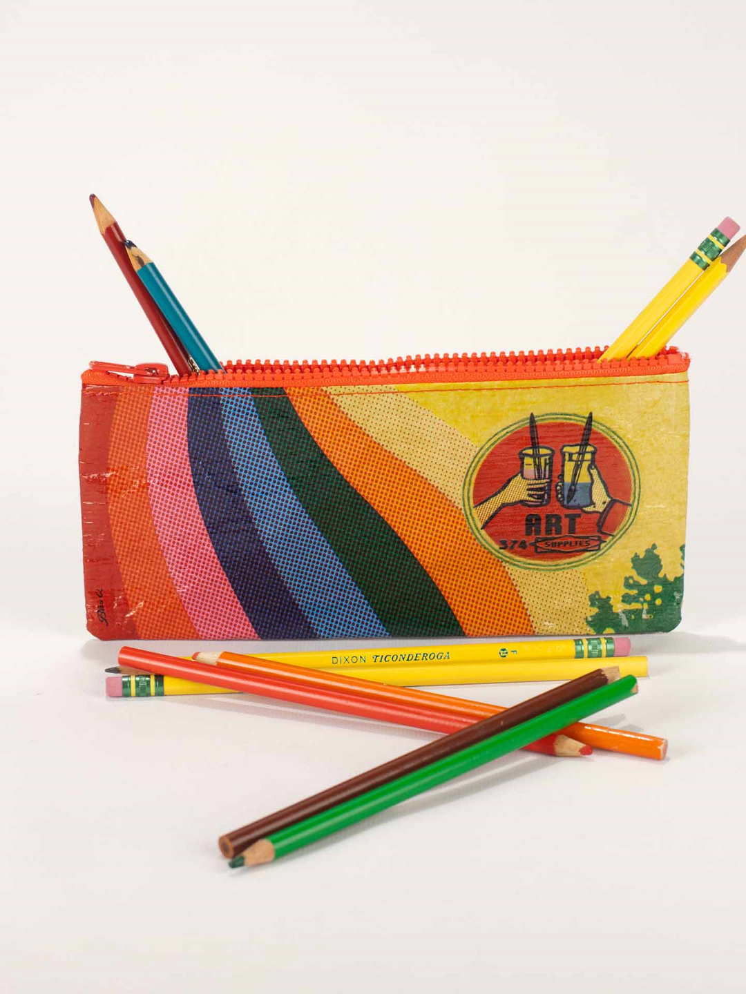 The Blue Q Art Supplies Pencil Case, crafted from recycled material, showcases a vibrant rainbow design and is equipped with a red zipper. Filled with colored pencils—some neatly tucked inside while others spill out onto the white surface—this conveniently-sized case is perfect for creating a mobile art studio on the go.