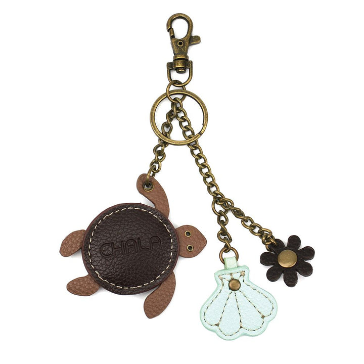 Introducing the Turtle Keychain by Chala: a delightful mini keychain with a bronze clasp, showcasing three charming accessories. It features a brown faux leather sea turtle with the word “CHALA” stitched on its shell, accompanied by a white seashell and a dark brown flower charm. All charms are elegantly connected by bronze chains.