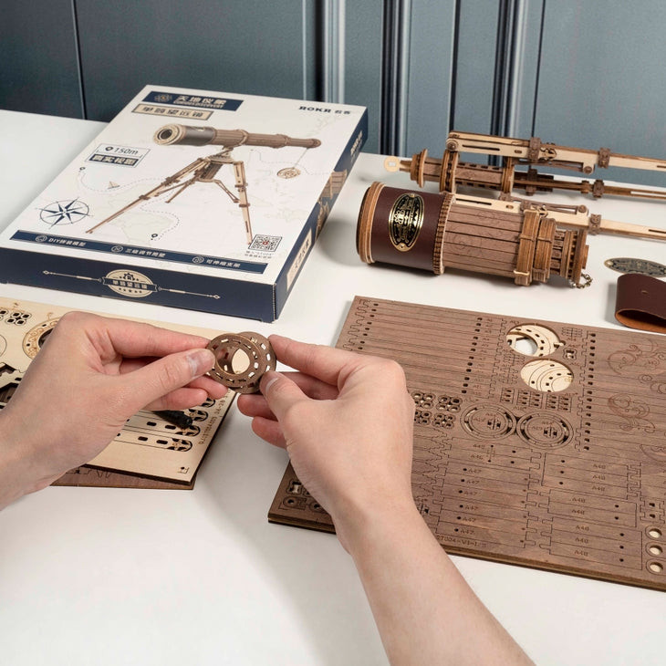 Two hands are working on assembling the DIY Wooden Puzzle: Monocular Telescope by Hands Craft, with multiple wooden pieces spread out on the table. The partially assembled monocular telescope and its packaging, which highlights an extendable telescopic lens in a pirate theme, can be seen in the background. The scene resembles a hobby or crafting space.