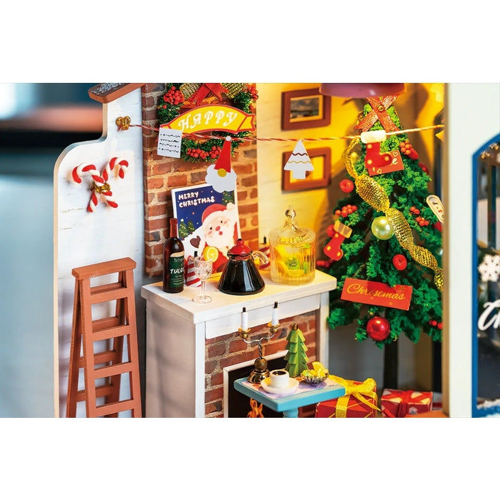 A cozy Christmas scene features a decorated tree, colorful lights, a "Merry Christmas" sign, and gifts. A small table offers holiday treats beside a ladder set against a brick wall adorned with festive cards and candy canes. Nearby, the Robotime Christmas Patio DIY Miniature House Kit adds whimsical charm to the ambiance.