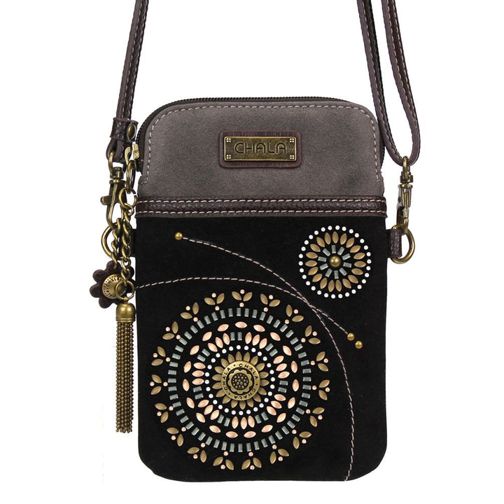 The Cell Phone XBody Bohemian Black by Chala is a crossbody bag that showcases a decorative Bohemian pattern with golden and bronze circular designs, floral motifs, and a brass tassel charm. Crafted from faux suede leather, the bag features a dark base with a brown top section and comes with an adjustable strap for personalized comfort.