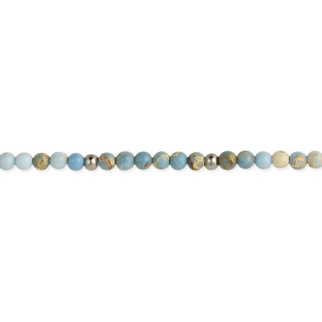 The Blue Jasper Bead Anklet by ZAD Jewelry features polished, round beads in various shades of blue, aqua, and green, interspersed with silver plate beads and sleek Imperial Jasper. The arrangement, resembling a delicate anklet, lies in a straight line against the white background.