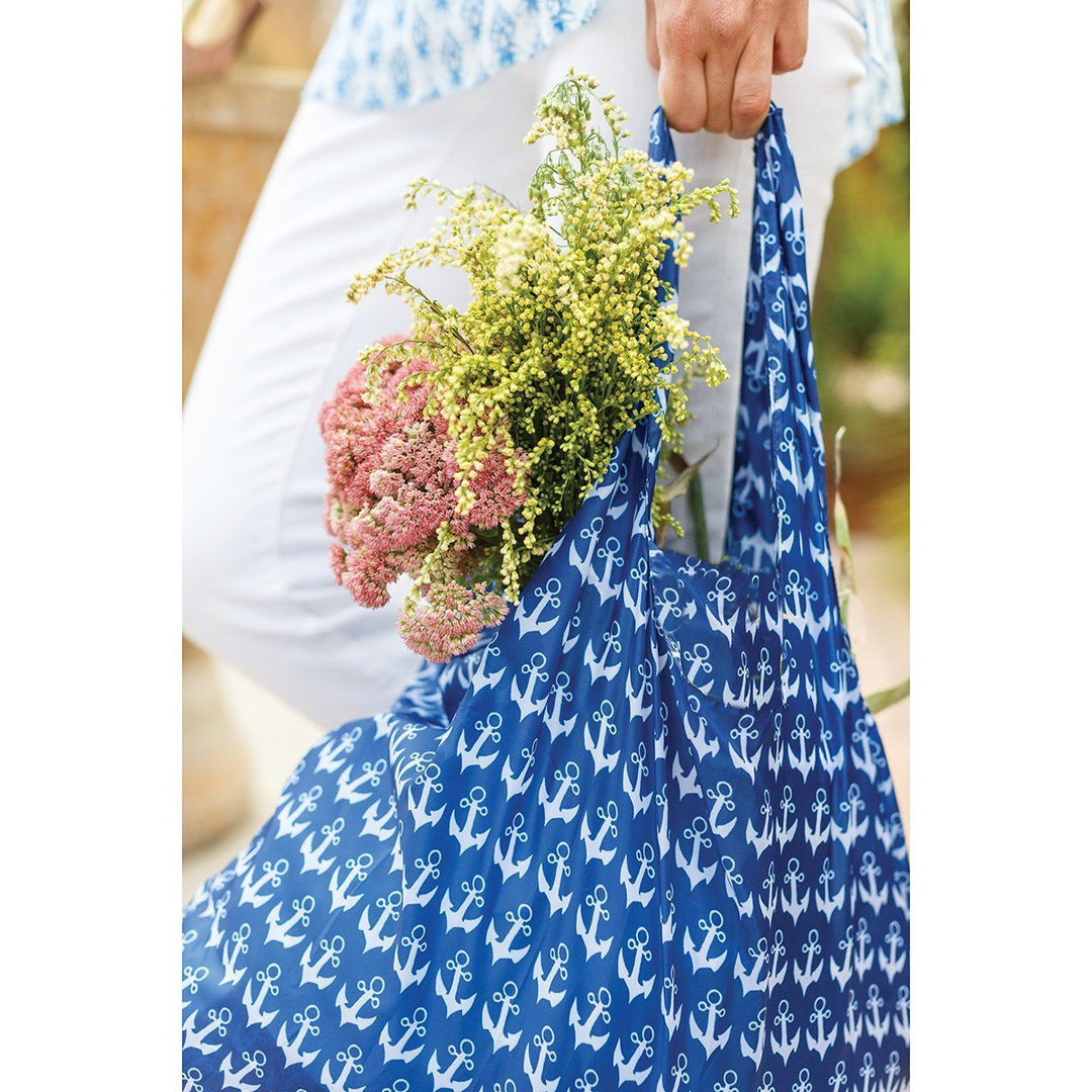 A person carrying a zzzRockFlowerPaper Anchor Navy Reusable Bag filled with yellow and pink flowers. The eco-friendly accessory complements their white pants and patterned blue shirt, showcasing a harmonious blend of style and sustainability.