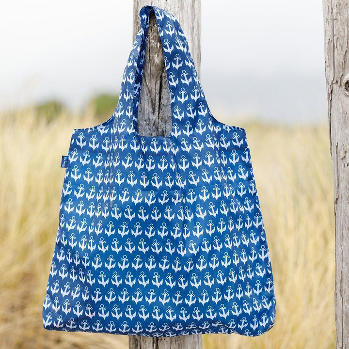 An Anchor Navy Reusable Bag from zzzRockFlowerPaper, featuring an eco-friendly blue print, hangs on a wooden post against a backdrop of blurred dry grass and a cloudy sky, evoking an outdoor setting.