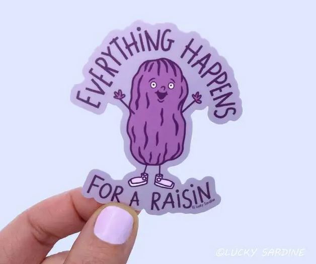 A hand with purple nail polish is holding the "Everything Happens For a Raisin" sticker, which features a cartoon raisin character with a happy face and raised arms. Made of durable vinyl, the sticker has the credit "Lucky Sardine" at the bottom right corner.