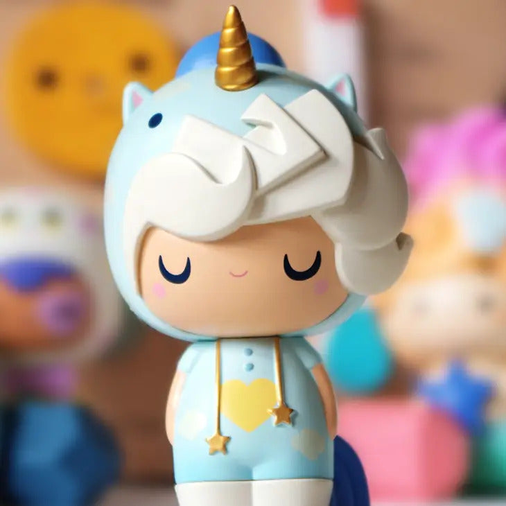 A cute, smiling figure wearing a light blue unicorn guardian costume with a golden horn is shown. The Little Starlight Girl vinyl doll by Momiji has blonde hair, pink cheeks, and is adorned with star-shaped accessories. The background is blurred with colorful shapes, making this Momiji icon truly enchanting.