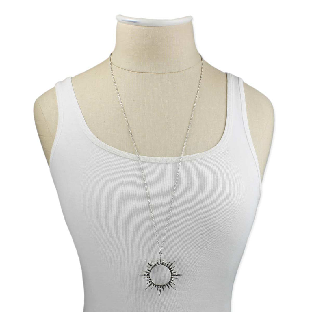 A mannequin torso dressed in a plain white tank top displays the Silver Sunburst Crystal Necklace by ZAD Jewelry. The fine link silver chain holds a pendant featuring a circular center surrounded by pointed rays.