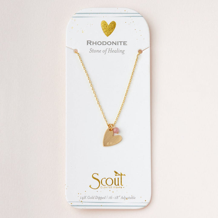 A delicate 14K gold necklace, named the Intention Neck Rhodonite/Gold from Scout Jewelry, is displayed on white packaging adorned with gold accents. The necklace features a heart-shaped pendant inlaid with semi-precious rhodonite gemstones. The packaging reads "Rhodonite - Stone of Healing." The adjustable chain's length ranges from 16 to 18 inches, and the brand name "Scout" is featured at the bottom.