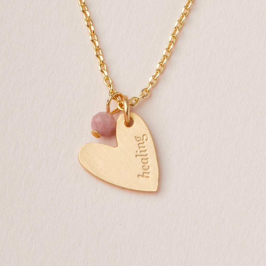 The Intention Neck Rhodonite/Gold by Scout Jewelry is a 14K gold necklace with a heart-shaped pendant and a small pink bead. The pendant is engraved with the word "healing." Featuring an adjustable chain, the necklace is displayed on a plain off-white background.