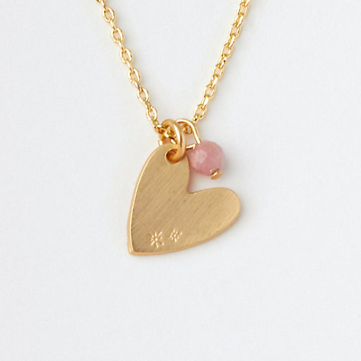 The Intention Neck Rhodonite/Gold by Scout Jewelry is a delicate 14K gold necklace that features a heart-shaped pendant adorned with subtle engravings. A small pink rhodonite gemstone is attached next to the heart on the same loop, hanging from a thin, adjustable gold chain, all set beautifully against a plain white background.