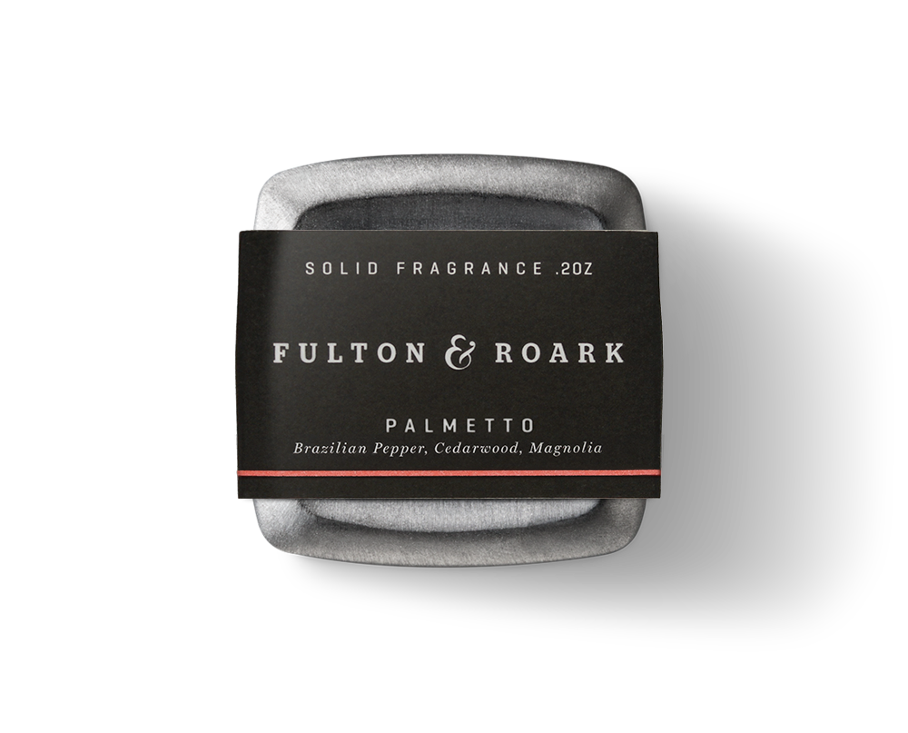 A sleek, metallic black container with rounded edges, labeled "Fulton & Roark," holds the Palmetto .2oz solid fragrance. This fragrance features notes of Brazilian Pepper, Cedarwood, and Magnolia.