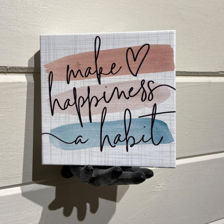 The "Make Happiness" sign by SincereSurroundings is a petite, handcrafted piece that showcases the phrase "make happiness a habit" in elegant cursive, with horizontal pink and blue brushstrokes. Ideal for shelf displays, it is set off beautifully by a dark, sculpted hand against a light-colored textured wall backdrop.