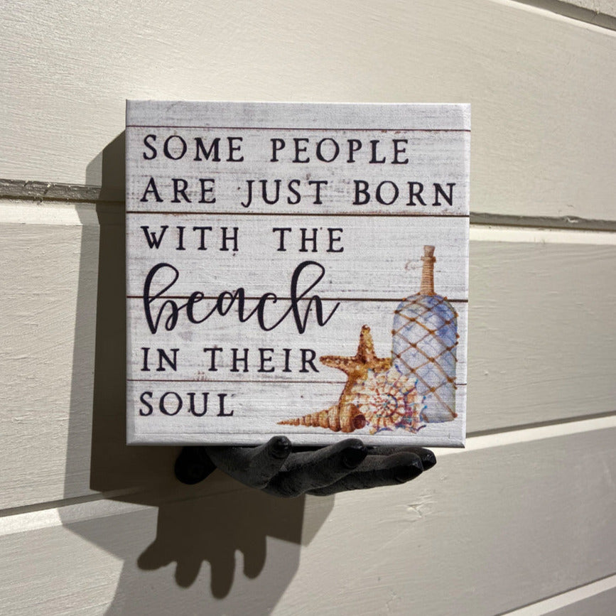 A hand holds a handcrafted wooden sign from SincereSurroundings called "Some People," featuring the text "Some people are just born with the beach in their soul." The sign is decorated with starfish, seashells, and a bottle, and is easily displayed as it rests against an off-white wall with horizontal lines.