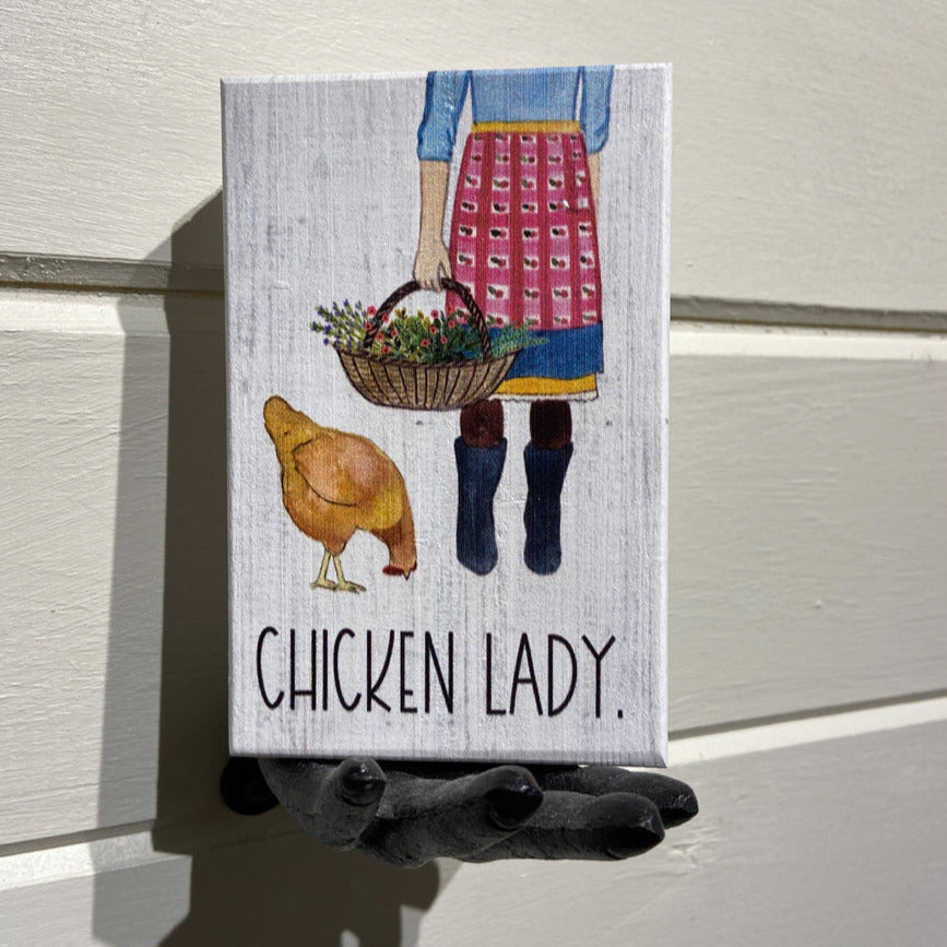 A handcrafted canvas sign from SincereSurroundings, titled "Chicken Lady," features a person holding a basket of flowers with a chicken beside them. The individual is dressed in a red apron, blue shirt, and black boots. Below the image, the text reads "Chicken Lady." The sign is held by a black mannequin hand for easy display, making it perfect to sit on your favorite shelf.