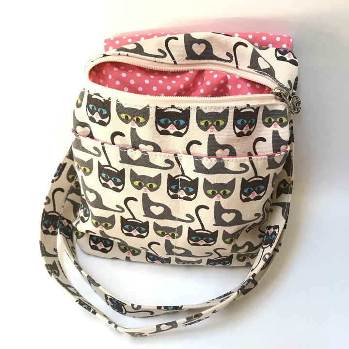 The "Sm Messenger Cat" crossbody bag by Bungalow 360 features a whimsical fabric with cat illustrations wearing glasses in blue, yellow, and pink. Crafted from 100% vegan materials, this unique small bag includes a pink polka dot lining at the top, a secure zipper closure, and an adjustable strap for convenience.