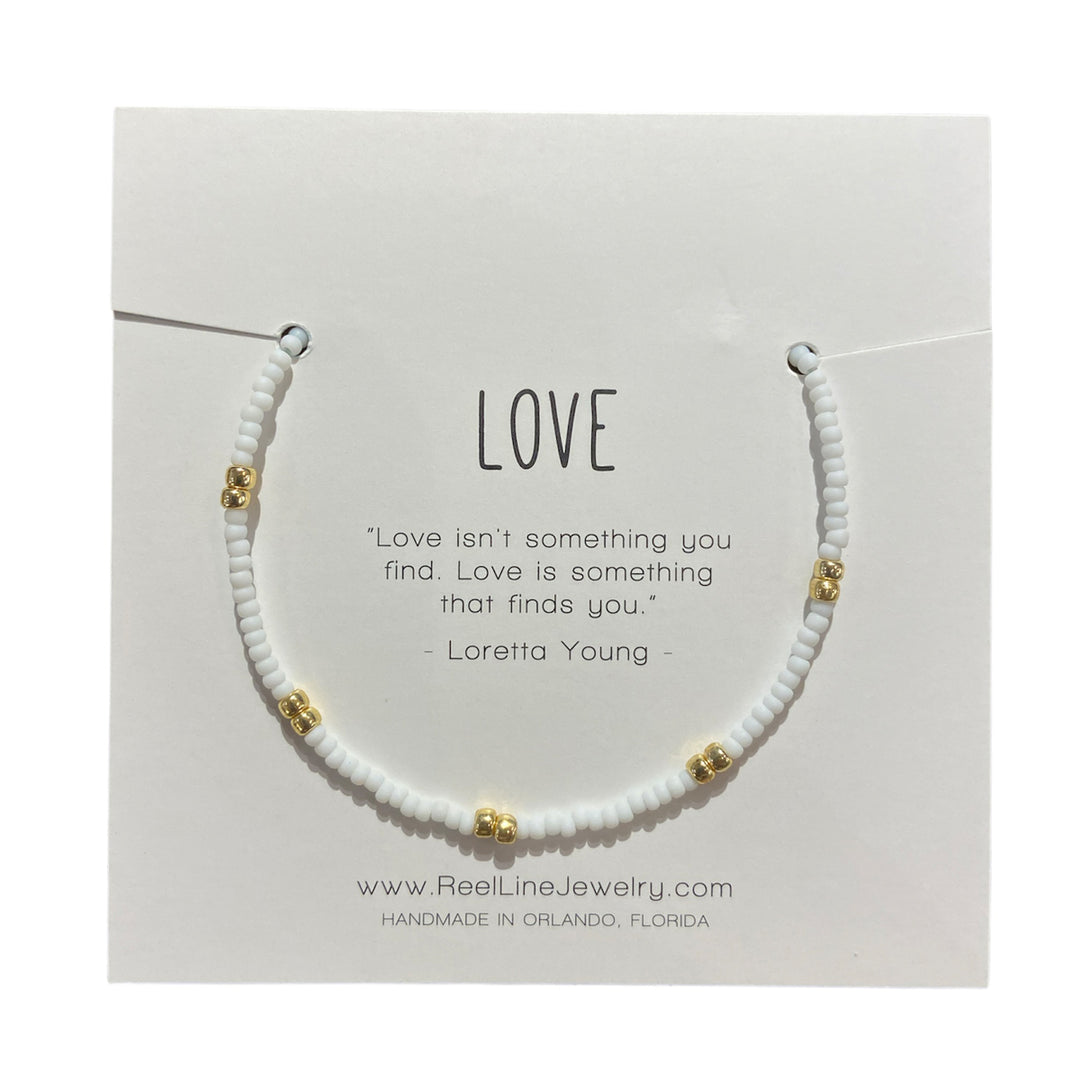 The "Love - Frost and Gold" bracelet by Reel Line Jewelry is an elegant glass beaded piece with white and gold beads, presented on a card. The card includes a quote from Loretta Young: "Love isn't something you find. Love is something that finds you." This adjustable handcrafted bracelet proudly states: "Handmade in Orlando, Florida.