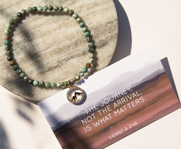 The Wanderlust Mountain Bracelet, adorned with African turquoise gemstones and a round pendant, is elegantly displayed on a stone surface. Positioned beneath it is a card with a gradient background showcasing the words: "The journey, not the arrival, is what matters. Lenny & Eva.