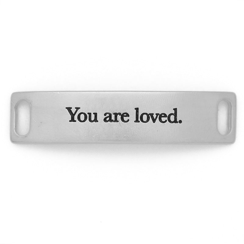 Introducing the ″You are loved - Silver″ by LENNY & EVA, this rectangular tag beautifully engraved with "You are loved" is designed in silver. It boasts a classic white background and comes with dual slots on each side, perfect for convenient attachment to bracelets or other accessories.