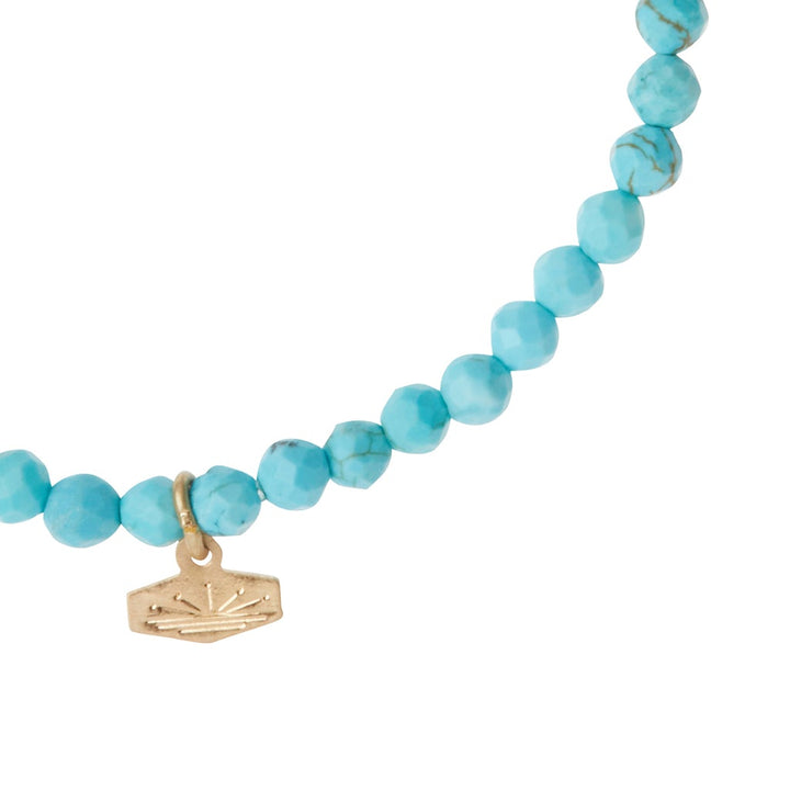 Introducing the Mini Bracelet Turquoise/Gold by Scout Jewelry: a beaded bracelet crafted with vibrant, faceted turquoise stone beads, adorned with a small, rectangular gold charm featuring an etched sunburst design. The stretch cord enhances its delicate and elegant appearance.