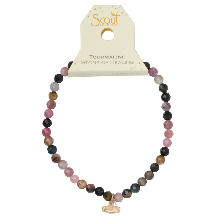 Introducing the Mini Bracelet Tourmaline/Gold from Scout Jewelry: A vibrant bracelet crafted with semi-precious tourmaline stones elegantly strung on a flexible stretch cord. It is presented on a card marked "Scout" and “Tourmaline Stone of Healing,” showcasing a delicate small gold charm with an intricate geometric design.