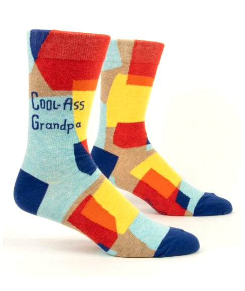 Introducing the Cool Ass Grampa Socks by Blue Q! These vibrant socks showcase a playful patchwork of red, yellow, blue, and brown rectangles set against a light blue background. Crafted from combed cotton with a hint of spandex for added comfort and flexibility, they boast blue toes, heels, and cuffs. The phrase "Cool-Ass Grandpa" is stylishly written on the side of one sock.