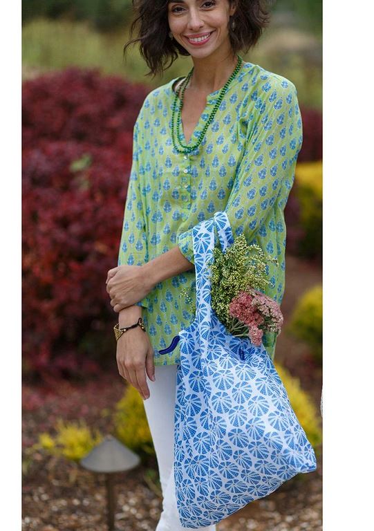 A person stands outdoors smiling, wearing a green patterned tunic and holding the eco-friendly Sea Urchin Reusable Bag by zzzRockFlowerPaper, which features floral patterns in blue. The reusable bag perfectly complements the vibrant red foliage and lush greenery in the background.