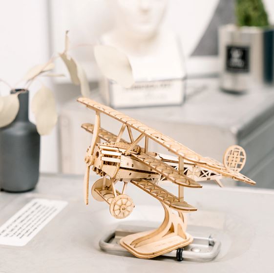 A Hands Craft DIY Airplane, a wooden model designed as a 3D puzzle, sits elegantly on a minimalist desk. In the background, various objects enhance the scene's aesthetic appeal, including a black vase filled with neutral-colored leaves and a couple of blurred decorative items.