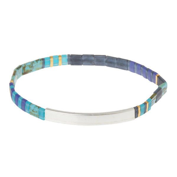 The Miyuki Brave Cobalt Silver bracelet by Scout Jewelry is a vibrant, slim accessory crafted from small, square Japanese glass Miyuki beads in varying shades of blue, purple, turquoise, and gold. It features a central flat, shiny metal segment engraved with empowering messages and is shaped into a circle.