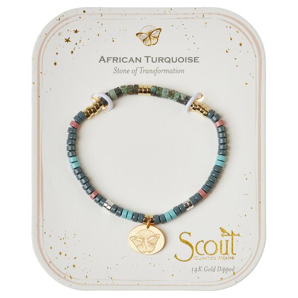 The Intention Charm African Turq/Gold bracelet by Scout Jewelry features semi-precious stones in shades of turquoise, grey, and pink, with gold accents and a gold pendant that has an embossed design. This stretch cord bracelet is packaged on a card that reads "African Turquoise: Stone of Transformation" and includes the branding "Scout Curated Wears".