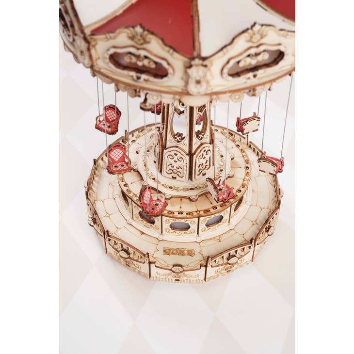 Introducing the Electro-Mechanical Wooden Puzzle: Swing Ride by Hands Craft, a meticulously detailed model of a vintage amusement park swing ride. This beautifully designed puzzle features ornate carvings and hanging swing seats, adorned in a harmonious mix of cream and deep red colors. The intricately crafted red swing seats are suspended from a multi-tiered, elaborately decorated base, capturing the essence of classic amusement park attractions.