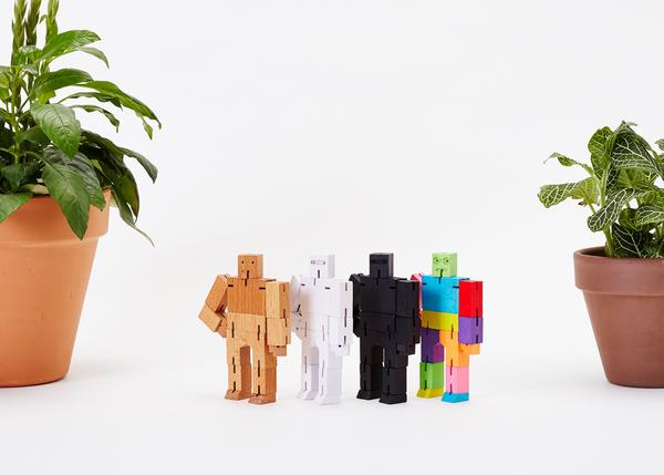 Arranged between two potted plants, four small blocky robot figures evoke the style of Japanese Shinto Kumi-ki puzzles. The robots—identified as Cubebot Micro Green Multi by Areaware—are colored light brown, white, black, and multicolored and stand against a plain white background.