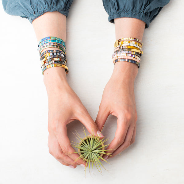 A person's hands holding a small air plant. The person is wearing a dark blue-green long-sleeved shirt, with both arms adorned with multiple colorful Scout Jewelry tile bracelets featuring Miyuki Brave Cobalt Silver beads. The background is a plain white surface.